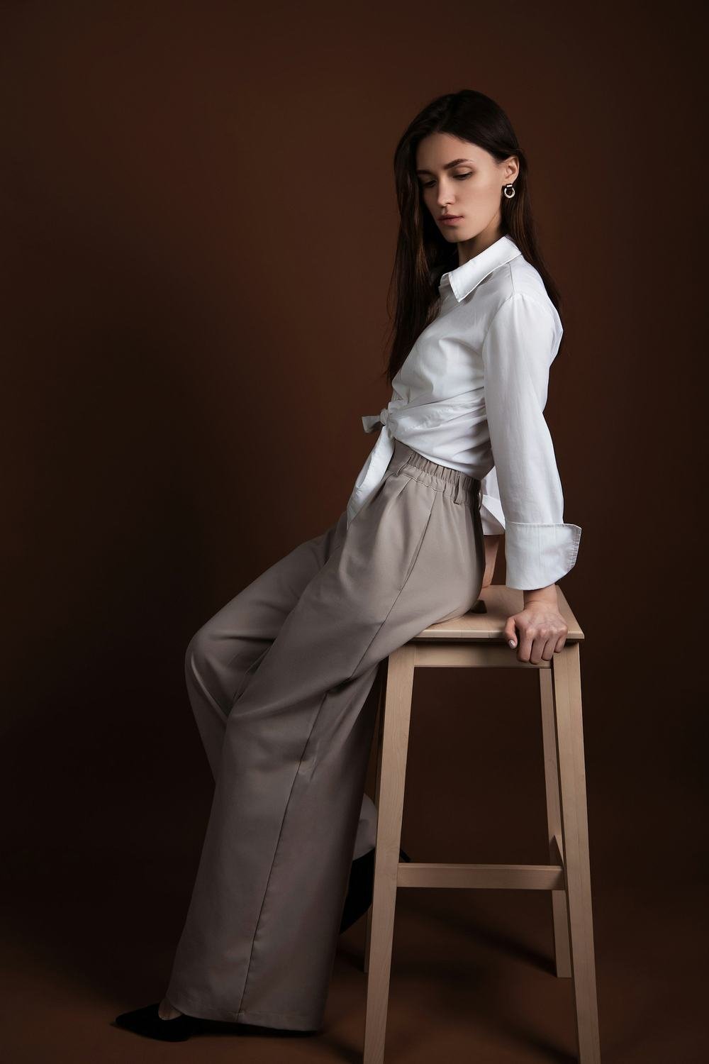 Modelo sentada em banco de madeira usa camisa branca de linho e calça pantalona areia